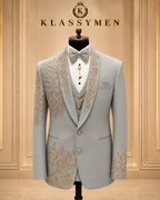 A light gray three-piece suit with ornate gold embroidery on the lapels, shoulders, and lower front, displayed on a mannequin with a matching gray patterned bow tie and white shirt. The background is a warm beige paneled wall with the brand name 'KLASSYMEN' displayed at the top.