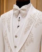 A close-up of a white formal tuxedo jacket on a mannequin, featuring intricate floral lace embroidery along the lapels and shoulders, paired with a matching white lace bow tie and a white textured vest.